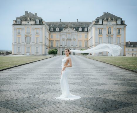 Braut in eleganter Kleider steht vor einem historischen Schloss im Freien.
