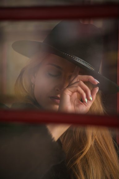 Frau mit langem Haar und Hut, die nachdenklich durch ein Fenster schaut.