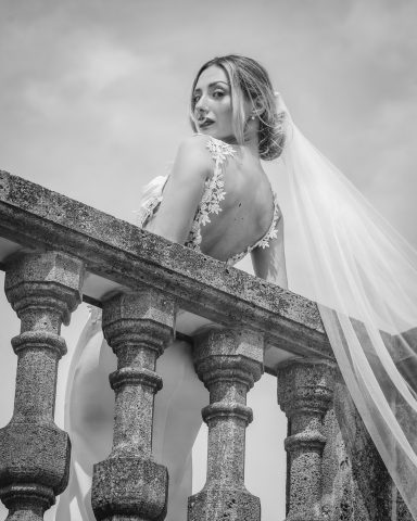 Braut in elegantem Kleid mit Schleier, schaut über eine Steinbalustrade zurück.